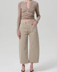 A person wears a fitted beige buttoned long-sleeve top, Citizens Of Humanity/AGOLDE Ayla Raw Hem Crop high-waisted wide-leg beige pinstripe pants in heavyweight cotton, and black heeled sandals, standing against a plain light background.