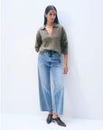 A woman with long straight dark hair and a neutral expression stands against a plain light background, wearing the White + Warren Cashmere Polo Top in brown, high-waisted wide-leg blue jeans, and pointed dark brown shoes.