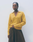 A woman with short braided hair and small hoop earrings stands against a light background, wearing the White + Warren Cashmere Polo Top in mustard yellow with a collar and a dark pleated skirt.