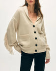 A woman models the White + Warren Cashmere Blend Luxe Fringe Coatigan, a cream-colored piece with black buttons, front pockets, and long fringed sleeves, styled with black pants against a plain light background.