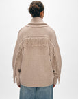 A person poses facing away in a White + Warren Cashmere Blend Luxe Fringe Coatigan and blue denim jeans. The beige coatigan, crafted from a luxurious merino wool-cashmere blend, features fringe detailing on the back and sleeves. Their hair is tied up against a plain, light background.
