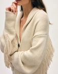 A woman wearing the White + Warren Cashmere Blend Luxe Fringe Coatigan with long fringed sleeves stands against a light background, her hand resting thoughtfully near her mouth and her face partially visible.