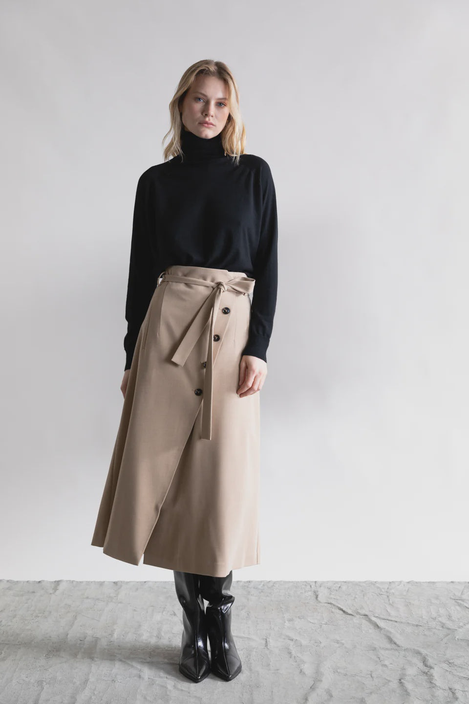 A woman stands against a white backdrop wearing the Psophia High Waisted Wrap Skirt, paired with a black turtleneck sweater and knee-high boots—an effortlessly chic look for the minimalist feminine wardrobe.