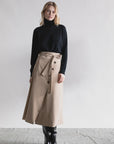 A woman stands against a white backdrop wearing the Psophia High Waisted Wrap Skirt, paired with a black turtleneck sweater and knee-high boots—an effortlessly chic look for the minimalist feminine wardrobe.