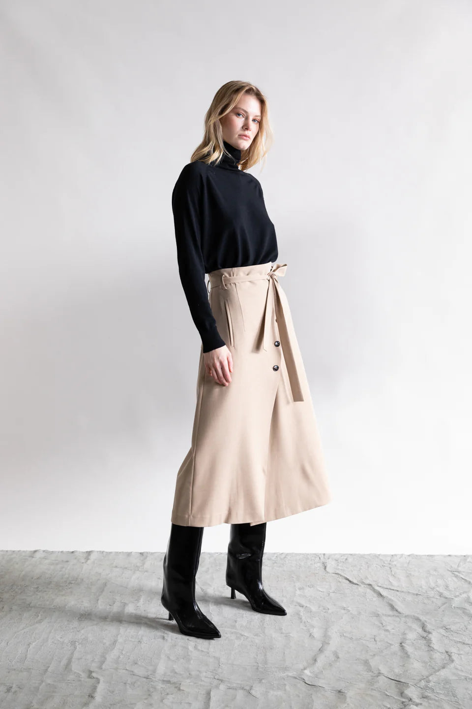 A woman stands before a plain background in a black turtleneck, Psophia High Waisted Wrap Skirt with button and tie accents, and knee-high boots—an ideal minimalist look.