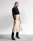 A woman stands before a plain background in a black turtleneck, Psophia High Waisted Wrap Skirt with button and tie accents, and knee-high boots—an ideal minimalist look.