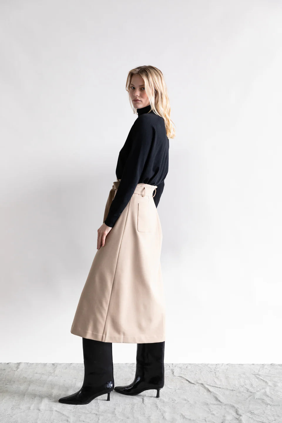 A woman with blonde, wavy hair and a neutral expression stands sideways against a white background, wearing a black long-sleeve top, Psophia's High Waisted Wrap Skirt in beige, and black knee-high boots—ideal for minimalist style.