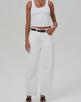 A person wears a white ribbed tank top and off-white high-waisted, relaxed-fit Miro jeans from Citizens of Humanity/AGOLDE with a black belt. They complete the look with black open-toe shoes and a necklace against a simple gray backdrop.
