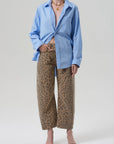 A person in the Miro shirt by Citizens Of Humanity/AGOLDE, loose light blue button-up, paired with off-white barrel leg jeans featuring black leopard print, black open-toe heels, and a gold necklace against a plain light backdrop.