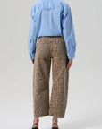 A person faces away, wearing the Miro off white barrel leg leopard print denim pants by Citizens Of Humanity/AGOLDE, paired with a light blue button-up shirt and black heeled sandals against a plain background.