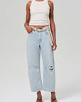 A person wearing the Miro White Sleeveless Top by Citizens Of Humanity/AGOLDE pairs it with light blue, non-stretch ripped denim jeans featuring a relaxed fit. They accessorize with a beige belt and brown sandals, standing against a simple light-colored background.