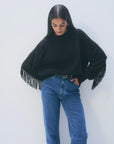 A woman with long dark hair wears the White + Warren Cashmere Luxe Fringe Crew and blue high-waisted jeans, standing against a plain light background and looking down with one hand in her pocket.