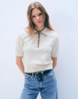 A woman with straight, light brown hair wears the White + Warren Cashmere Button Down Polo in cream, paired with blue jeans and a black belt. She stands against a plain white background, looking seriously at the camera.