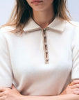 A woman wears the White + Warren Cashmere Button Down Polo in cream, featuring short sleeves, a collar, and fabric-covered front buttons. Her arms are crossed and she accessorizes with gold rings on her fingers.