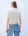 A person with long, light brown hair stands with their back to the camera against a plain light background, wearing a White + Warren Cashmere Button Down Polo in cream and light blue jeans.