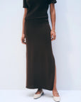 A person stands against a plain background wearing the Luxe Cotton Skirt by White + Warren in dark brown with a matching short-sleeve top featuring a side slit, styled with white pointed-toe flats and a delicate bracelet.