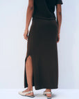 A person stands against a light background, shown from the back, wearing the White + Warren Luxe Cotton Skirt in dark brown with a side slit, paired with a matching top, gold bracelet, and beige slingback flats.