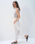A woman with wavy hair stands sideways against a plain background, wearing the White + Warren Cashmere Windowpane Vest, white high-waisted pocket pants, and black sandals. Her hands are in her pockets as she looks slightly down.