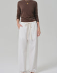 A person stands against a plain background wearing a brown long-sleeve shirt, Citizens Of Humanity/AGOLDE aurora pull on lightweight summer pants in an off-white cotton flax blend, black sandals, and layered gold necklaces.