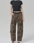 A person wears a fitted black crop top with Citizens Of Humanity/AGOLDE flight pant in camo/khaki, styled with a black belt, black sandals, and a gold bracelet, standing against a plain light gray background.