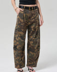 A person stands with one hand in a pocket against a plain background, wearing Citizens Of Humanity/AGOLDE flight pant in camo/Khaki, a black belt with gold buckle, black crop top, gold bracelet, and black heeled sandals.