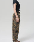 Person stands in profile against a plain background, wearing a fitted black short-sleeve top, Citizens Of Humanity/AGOLDE flight pant in camo/Khaki, a black belt, gold bracelet, and black heeled sandals.