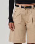 Dressed in a black long-sleeve top and Cara Pleated Shorts by Citizens Of Humanity/AGOLDE in beige lightweight twill, with a gold-buckled black belt enhancing the waist, this look seamlessly blends textures on the upper legs and visible arm.