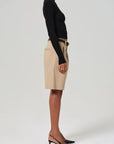A person wearing a black turtleneck and Citizens Of Humanity/AGOLDE's Cara Pleated Short in beige stands in profile, highlighting their lightweight twill outfit. They pair it with black slingback heels, hands relaxed by their sides against a plain, light backdrop.