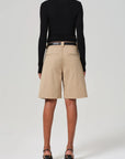 A person stands facing away, wearing a black turtleneck with Citizens Of Humanity/AGOLDE Cara Pleated Short in beige lightweight twill and black high-heeled sandals, set against a plain light gray background.