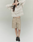A person wearing the Cara Pleated Short by Citizens Of Humanity/AGOLDE, a beige and white striped sweater, and black ankle boots stands against a plain background. With long hair, they playfully cover their mouth with the sweater's sleeve, giving off a vintage vibe.