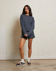 A woman with long dark hair poses casually indoors against a plain wall, wearing the Perfectwhitetee French Terry Sweatshirt and matching shorts in dark gray, paired with white socks and brown Nike sneakers.
