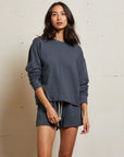 A woman with long dark hair and a neutral expression stands against a light-colored wall, wearing the Perfectwhitetee French Terry Sweatshirt with matching shorts featuring cream drawstrings.