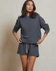 A woman stands against a plain light wall, wearing the Perfectwhitetee French Terry Sweatshirt and matching shorts with a white drawstring. She looks to the side with one hand on her hip and the other adjusting her sweatshirt.