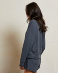 A woman with long dark hair stands sideways against a light wall, wearing the Perfectwhitetee French Terry Sweatshirt and matching dark gray shorts for an effortlessly casual look, her face partially turned from the camera.
