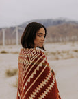 A woman with shoulder-length dark hair stands in a desert, wrapped in the Faire Achuar Blanket—red and beige, geometric-patterned, made from recycled acrylic. Wind turbines and hazy mountains form the background.