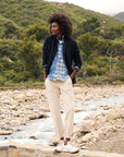 Clad in a Frank & Eileen Dublin Knit Blazer over a tailored-fit denim jacket and a 100% cotton dark sweater, a person stands on a rocky surface near a flowing river. Their hands rest in the pockets of beige pants, set against the backdrop of a lush, green mountainous landscape.