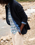 Standing by a rocky riverbank, a person is dressed in a sophisticated Dublin Knit Blazer from Frank & Eileen, layered over a blue plaid shirt. They pair this ensemble with beige pants, hands casually tucked into their pockets as they look downwards. Curly hair frames their face, enhancing the casual elegance of their style.