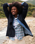 A woman stands beside a rocky, flowing river with lush green hills in the backdrop. She is smiling, wearing a plaid shirt and denim paired with the tailored Dublin Knit Blazer from Frank & Eileen. Her curly hair is loose as she raises her arms.