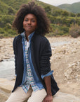 A person with curly hair is wearing a layered outfit, featuring a plaid shirt and the Dublin Knit Blazer by Frank & Eileen. They're seated on a stone wall by the rocky riverbank, surrounded by green hills and trees.