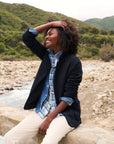 A person with curly hair and a joyful expression sits on a stone wall near a rocky riverbed, effortlessly rocking the Dublin Knit Blazer by Frank & Eileen over a plaid shirt and cream pants. Green hills and a cloudy sky serve as the perfect backdrop.
