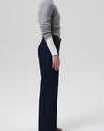 A person stands in profile against a plain background, wearing a fitted grey long-sleeve sweater, Citizens Of Humanity Annina Pocket Trouser in dark indigo denim, and black open-toe sandals.