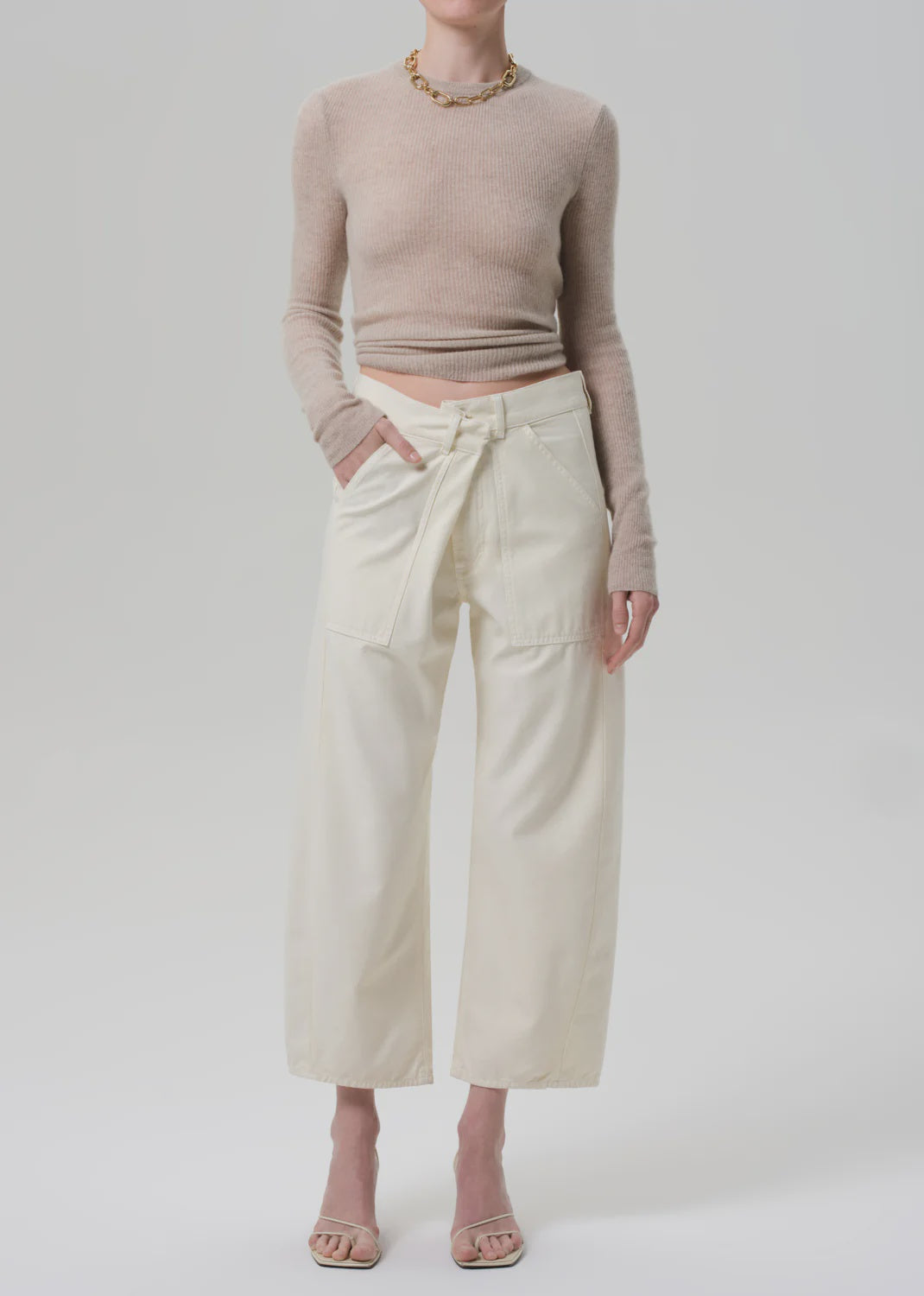 A person wears the Ines Crop Utility Trouser by Citizens Of Humanity/AGOLDE—high-rise, cropped, regenerative cotton twill with utility pockets—paired with a fitted long-sleeve beige crop top and open-toe heeled sandals against a plain background.