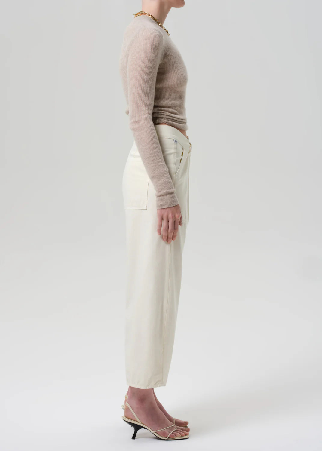 A person stands in profile against a plain background, wearing a fitted beige long-sleeve top and Citizens Of Humanity Ines Crop Utility Trouser, styled with strappy heeled sandals for a minimalist, casual look.