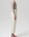 A person stands in profile against a plain background, wearing a fitted beige long-sleeve top and Citizens Of Humanity Ines Crop Utility Trouser, styled with strappy heeled sandals for a minimalist, casual look.