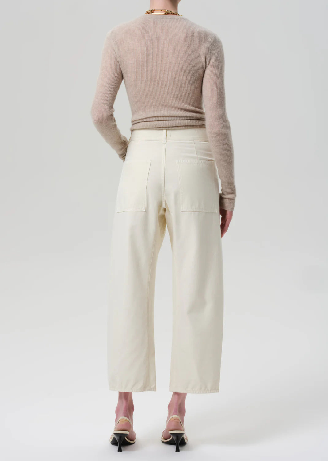 A person faces away wearing Citizens of Humanity's Ines Crop Utility Trouser, a high-rise cropped style in regenerative cotton twill, paired with a sheer beige long-sleeve top and black ankle-strap heeled sandals against a plain light background.
