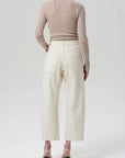 A person faces away wearing Citizens of Humanity's Ines Crop Utility Trouser, a high-rise cropped style in regenerative cotton twill, paired with a sheer beige long-sleeve top and black ankle-strap heeled sandals against a plain light background.