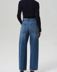 A person stands facing away from the camera, wearing a black long-sleeve top and Citizens Of Humanity/AGOLDE Ines Utility Trouser jeans in medium indigo denim with large back pockets, paired with black heeled sandals against a plain light background.
