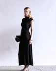 A woman with shoulder-length blonde hair stands against a plain background, wearing the Psophia Naia Drape Dress in black, gold shoes with socks, and holding a black clutch. Her expression is neutral.