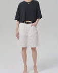A person wears the Citizens Of Humanity/AGOLDE court denim short in blanc with a loose black T-shirt, a black belt, open-toe sandals, and simple jewelry, standing against a plain light gray background.