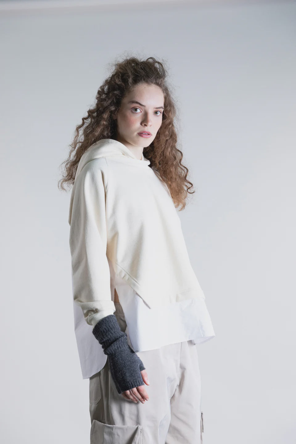 A woman with long curly hair wears the Psophia Cropped Hoodie W/ Shirt Hem and beige cargo pants, styled with dark fingerless gloves, standing against a plain light background.